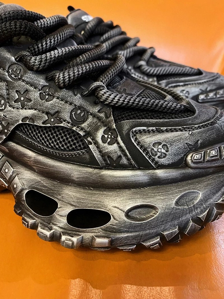 Low-angle close-up of a black techwear chunky sneaker showing intricate embossed patterns, rope laces, mesh inserts, and oval sole cutouts