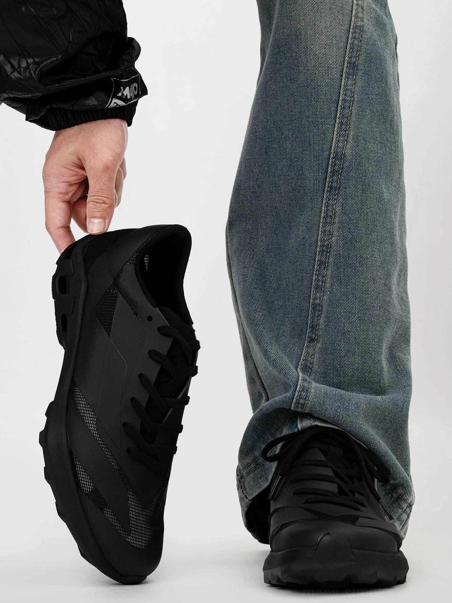 Black techwear trail sneakers shown from a rear oblique angle with a chunky anti-slip EVA outsole, matte mesh upper, and casual blue jeans streetwear styling
