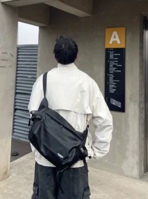 Black techwear utility crossbody sling bag worn across the back with white streetwear jacket and dark cargo pants in an industrial elevator lobby