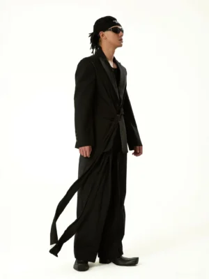 Side profile of a tailored black blazer coat showing the twisted waist drape, smooth suiting fabric, slim sleeves, and elongated avant-garde silhouette