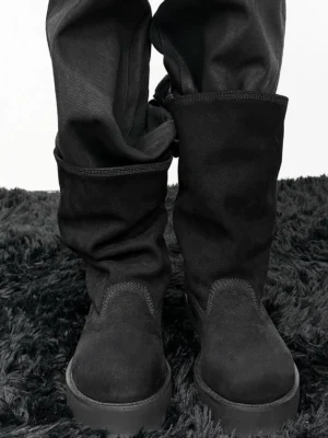 Model wearing black tumbled leather slouch mid-calf platform boots with straight black trousers on dark rug