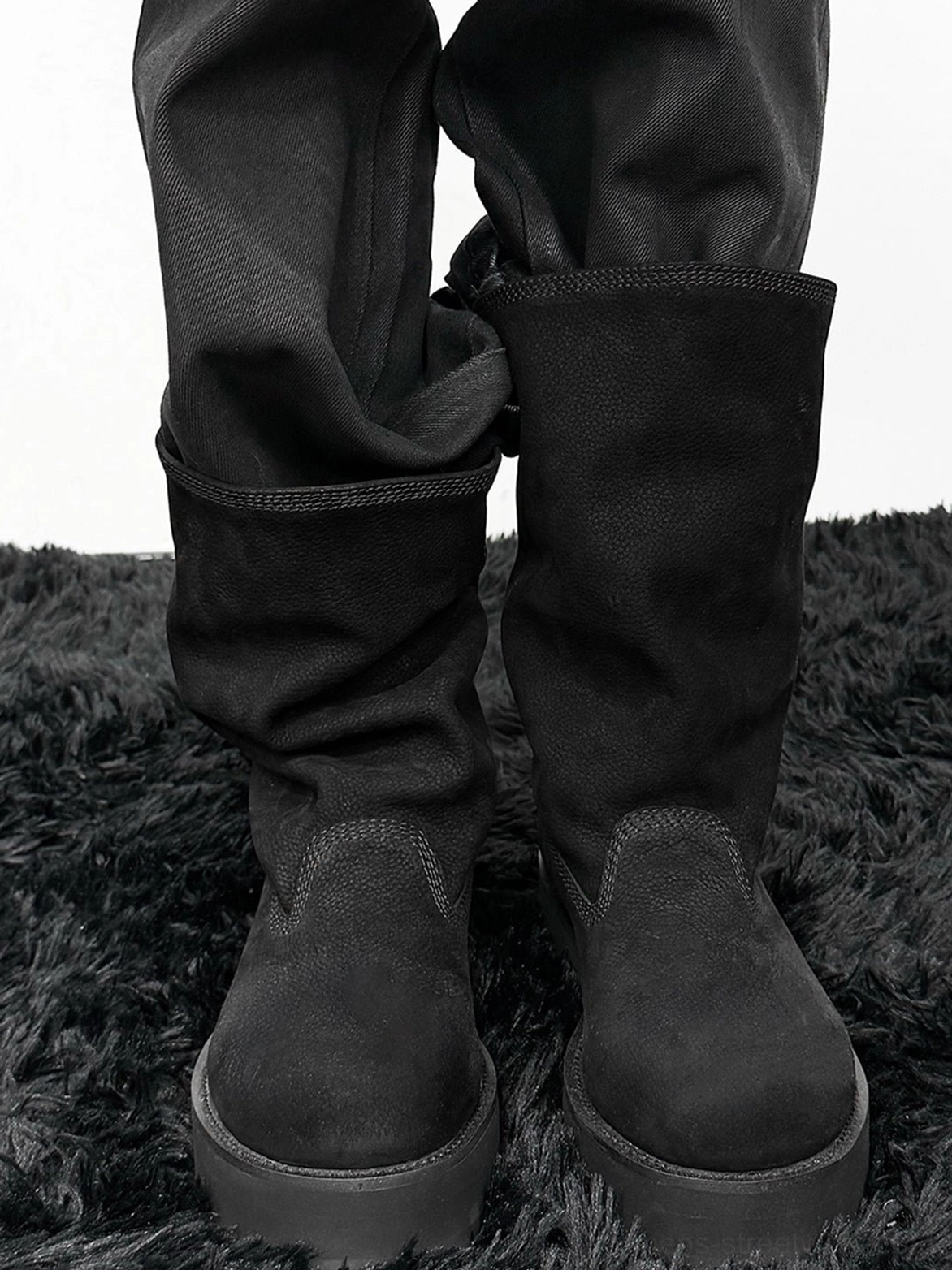 Model wearing black tumbled leather slouch mid-calf platform boots with straight black trousers on dark rug
