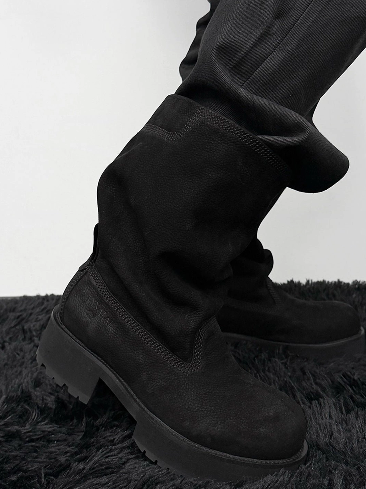 Side wearing view of black tumbled leather slouch mid-calf platform boots paired with loose black pants