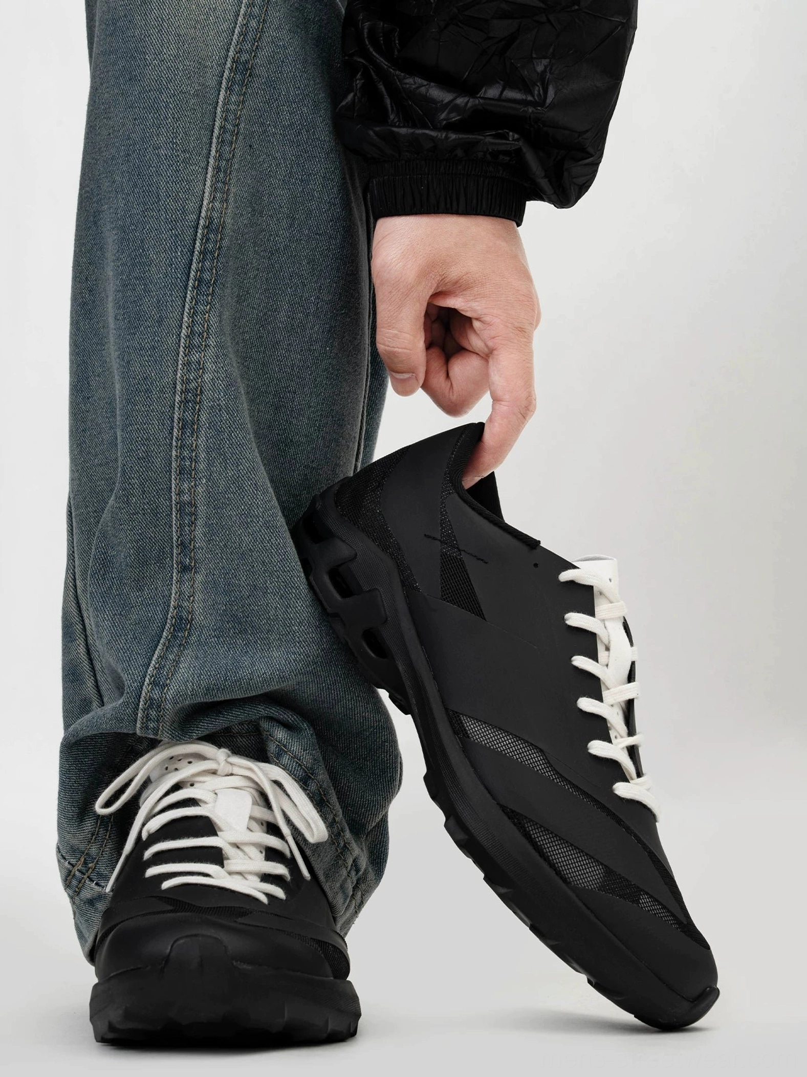 Black urban outdoor sneakers held at a rear side angle showing rugged wave traction outsole, matte upper, and white lace streetwear styling