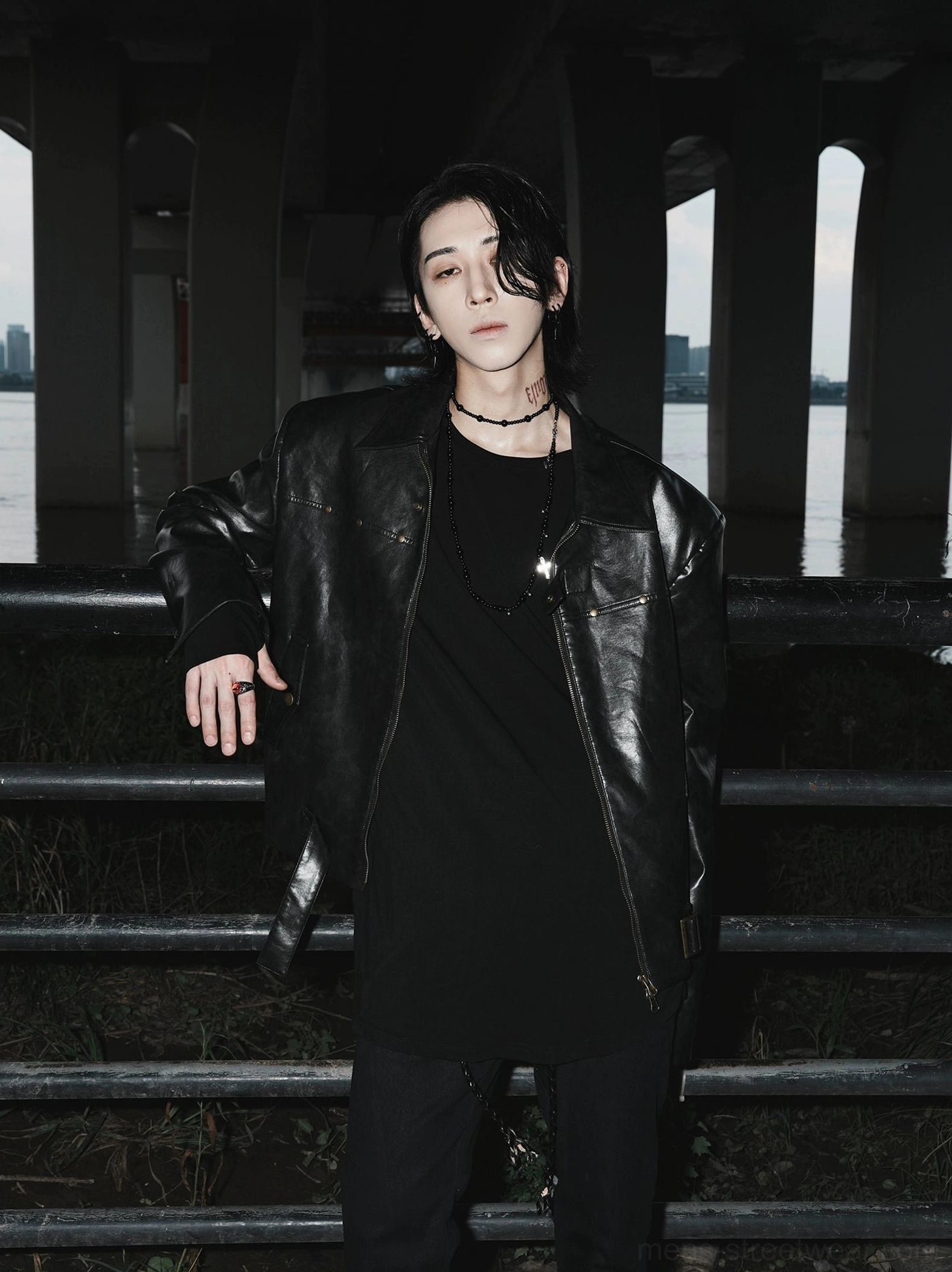 Black vintage faux leather motorcycle jacket styled with a black tee in an urban riverside setting, featuring bronze metal accents and relaxed streetwear fit