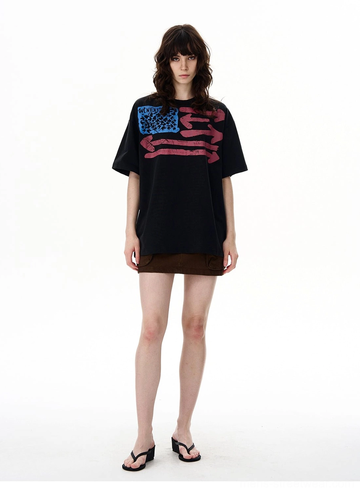 Female model wearing a black washed vintage flag graphic oversized T-shirt styled with a brown mini skirt and black flip flops