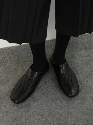 Black streamlined wave stitch leather slip-on loafers worn with black socks and a dark pleated skirt in a high-angle streetwear outfit view