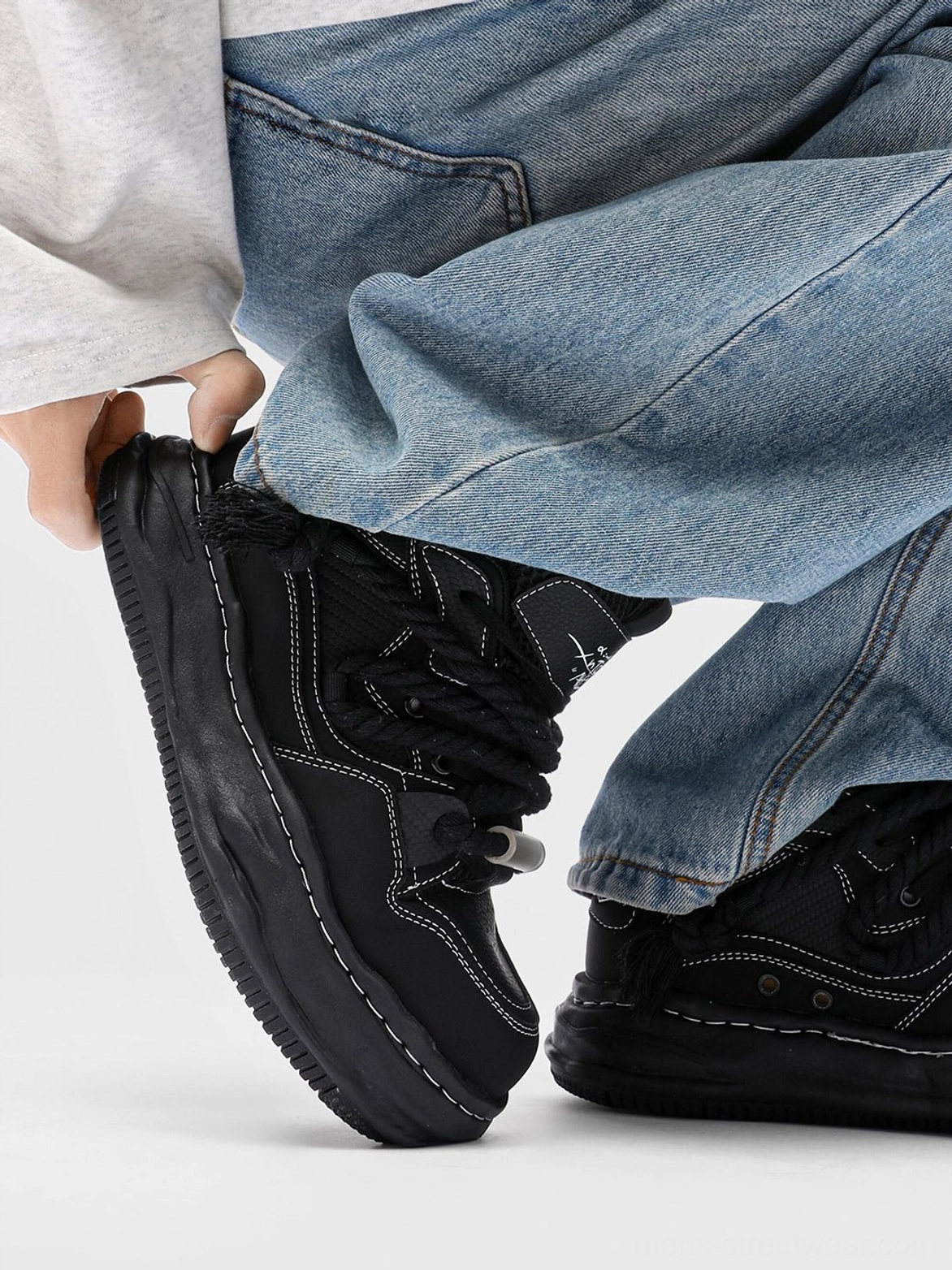 Black wavy platform sneaker held beside loose blue jeans, displaying the sculpted distorted outsole, rounded upper, and detailed contrast stitching