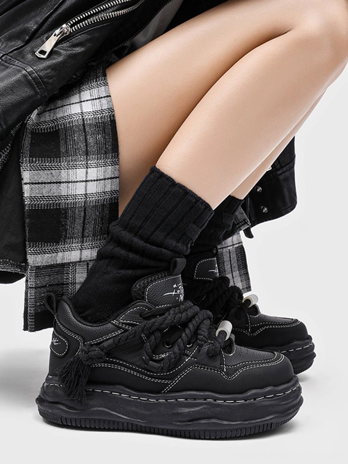 Close-up of black wavy platform sneakers worn with black crew socks and layered plaid and leather streetwear, showing the heel tassel and thick sole