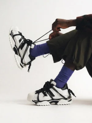 Black and white sculpted sole streetwear sneakers worn with dark green shorts and purple crew socks in a low angle studio view