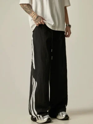 Side view of black wide-leg athletic track pants with contrasting white double side stripes, elastic drawstring waist, and relaxed streetwear fit