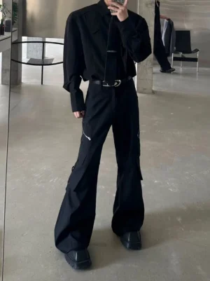 Black wide-leg cargo trousers shown from the front with zippered utility pockets, flared stacked hem, black belt, and chunky platform shoes in a minimalist concrete studio