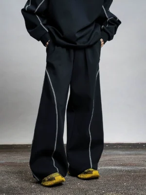 Black wide-leg contrast stitch sweatpants with oversized Korean streetwear fit, white curved topstitching, and casual sneakers in a minimalist studio