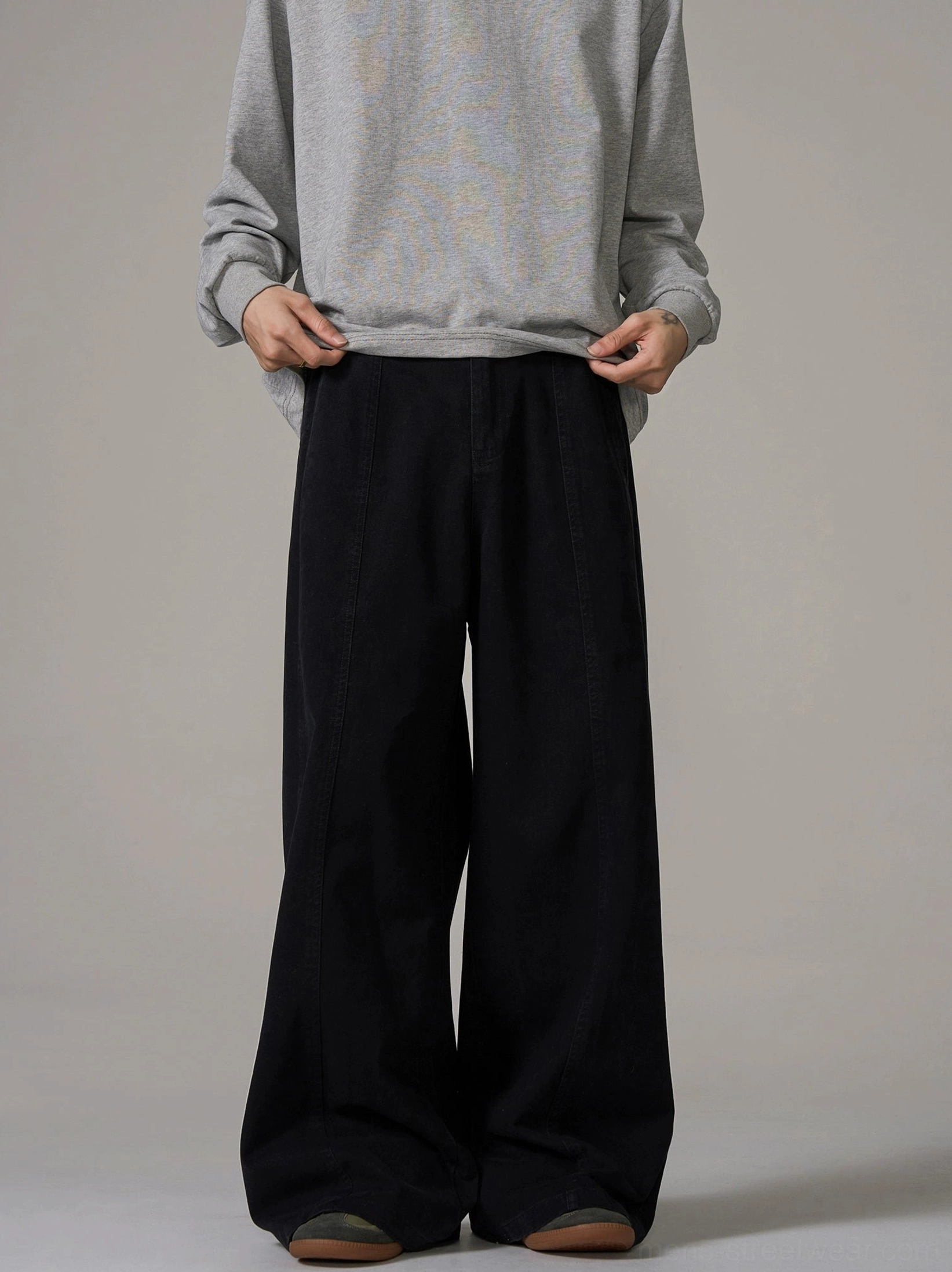 Male model wearing black wide-leg streetwear trousers with central front seam and relaxed fit against a plain grey studio background