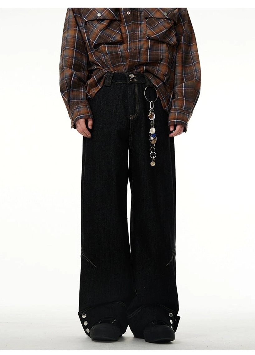 Front view of black wide-leg cuffed jeans with a decorative metal belt-loop pendant, rolled button hems, and layered casual streetwear styling