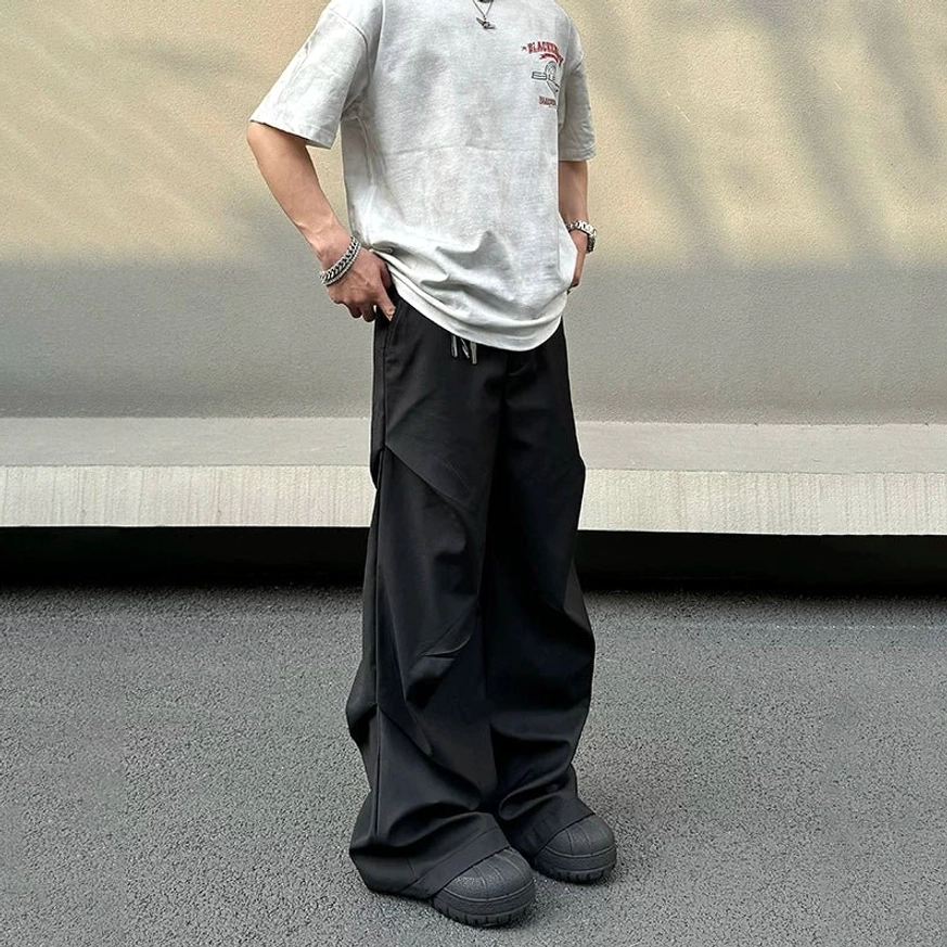 Black wide-leg draped trousers shown from a rear three-quarter angle with extra-long stacked legs, oversized light grey T-shirt, platform sneakers, and urban streetwear styling