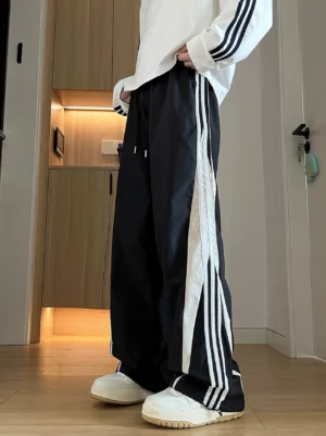Black wide-leg track pants with white triple stripe side panels shown from the front three-quarter view with white sneakers