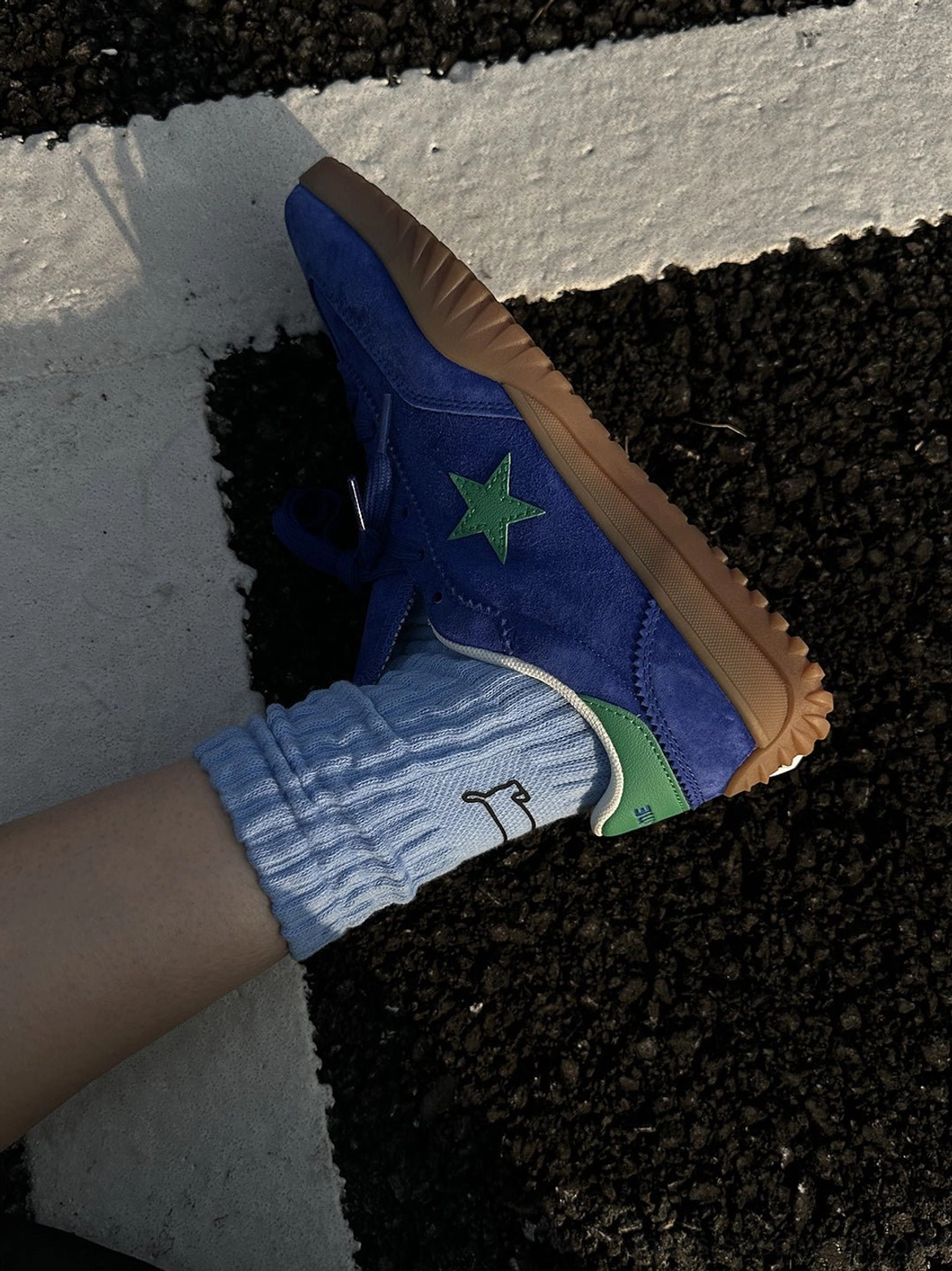Close-up of blue suede star trainers with green embroidery and heel tab worn with a ribbed light blue crew sock on an urban asphalt street
