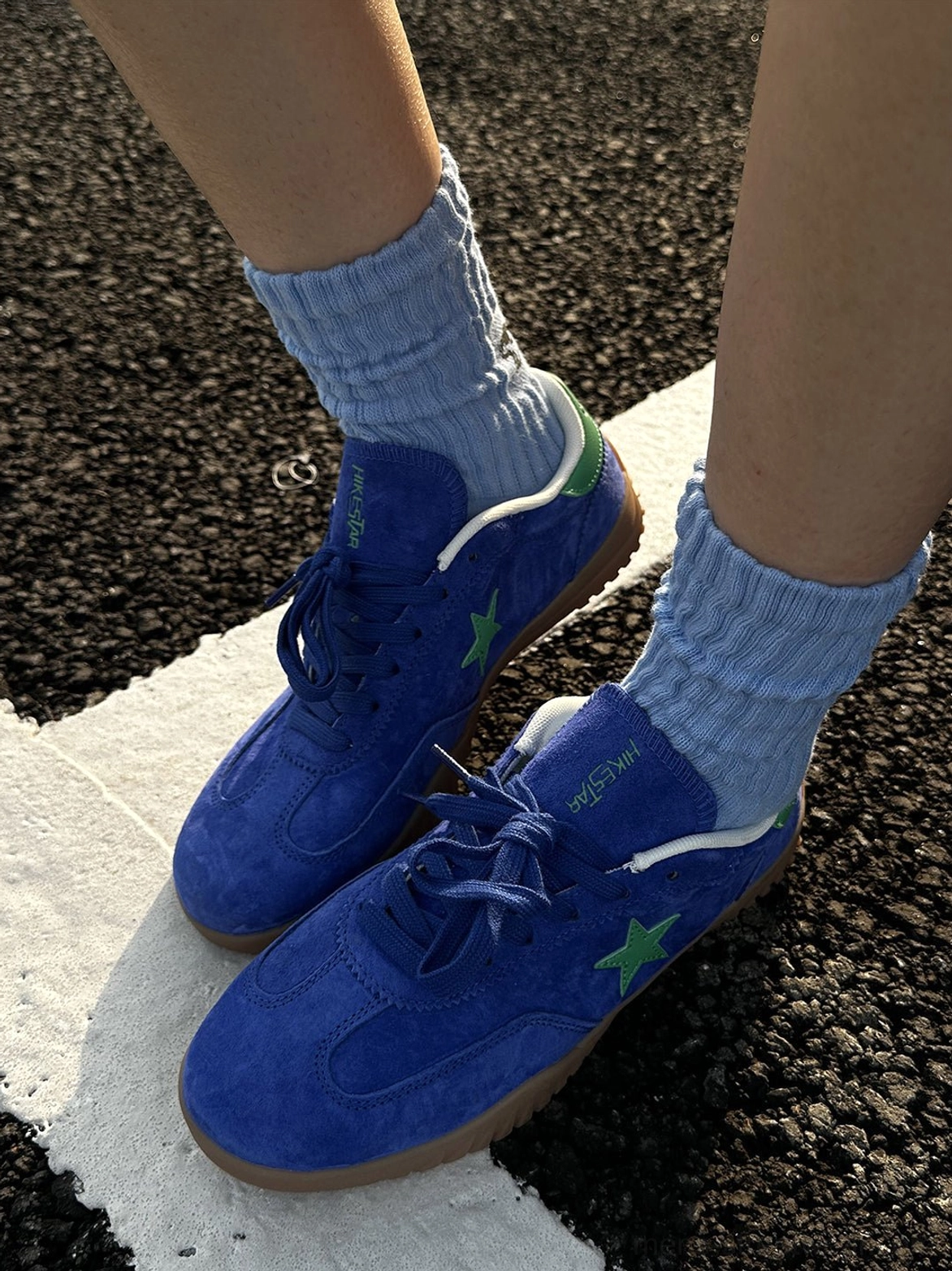 Low front side view of blue suede streetwear trainers showing the gum rubber outsole, green heel accents, and ribbed light blue crew socks