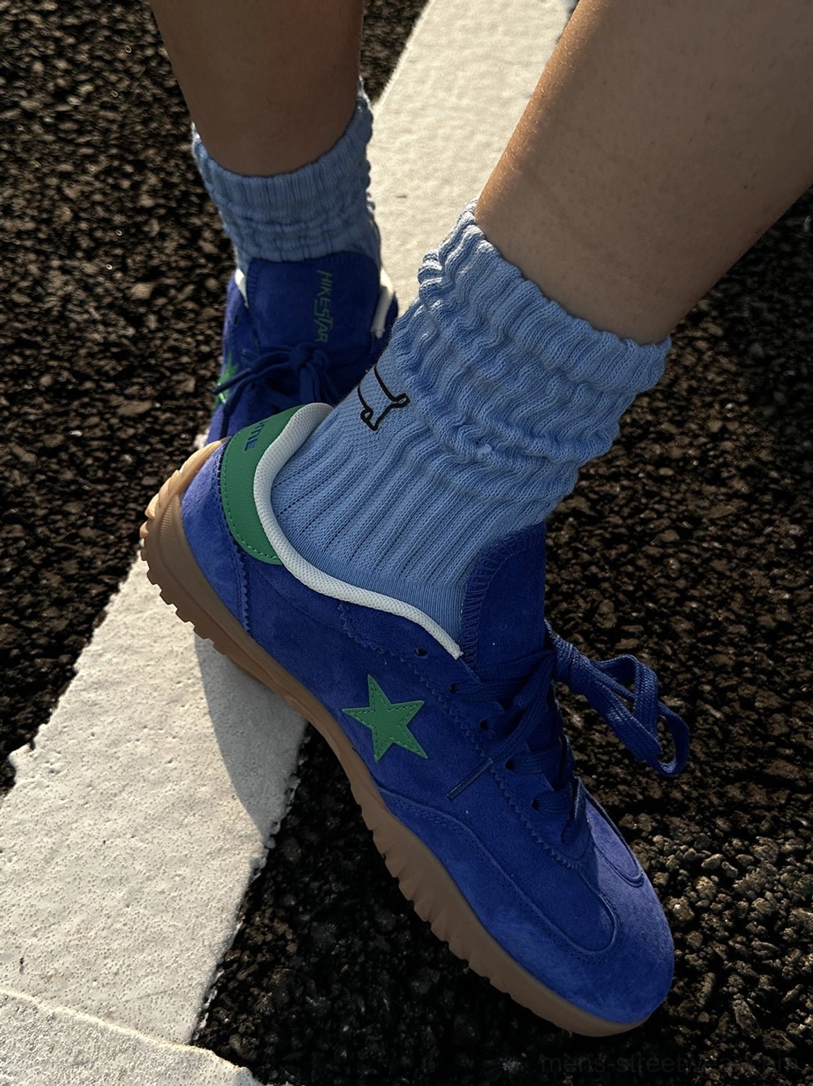 Close side view of one blue suede trainer featuring green star embroidery, green heel tab, gum sole, and ribbed light blue sock