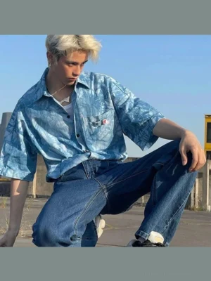 Male model wearing a blue tie-dye oversized short sleeve button-up streetwear shirt with denim jeans on an urban rooftop