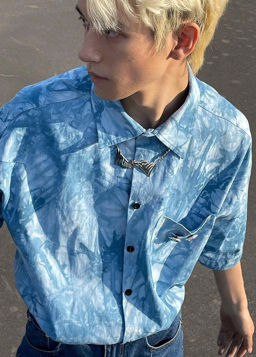 Upper close-up of a male model wearing a blue tie-dye shirt with pointed collar black buttons and geometric necklace