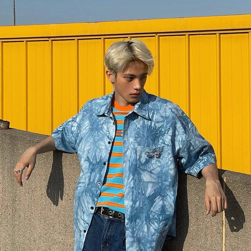 Front three-quarter view of a male model wearing a loose blue tie-dye shirt layered over a striped T-shirt