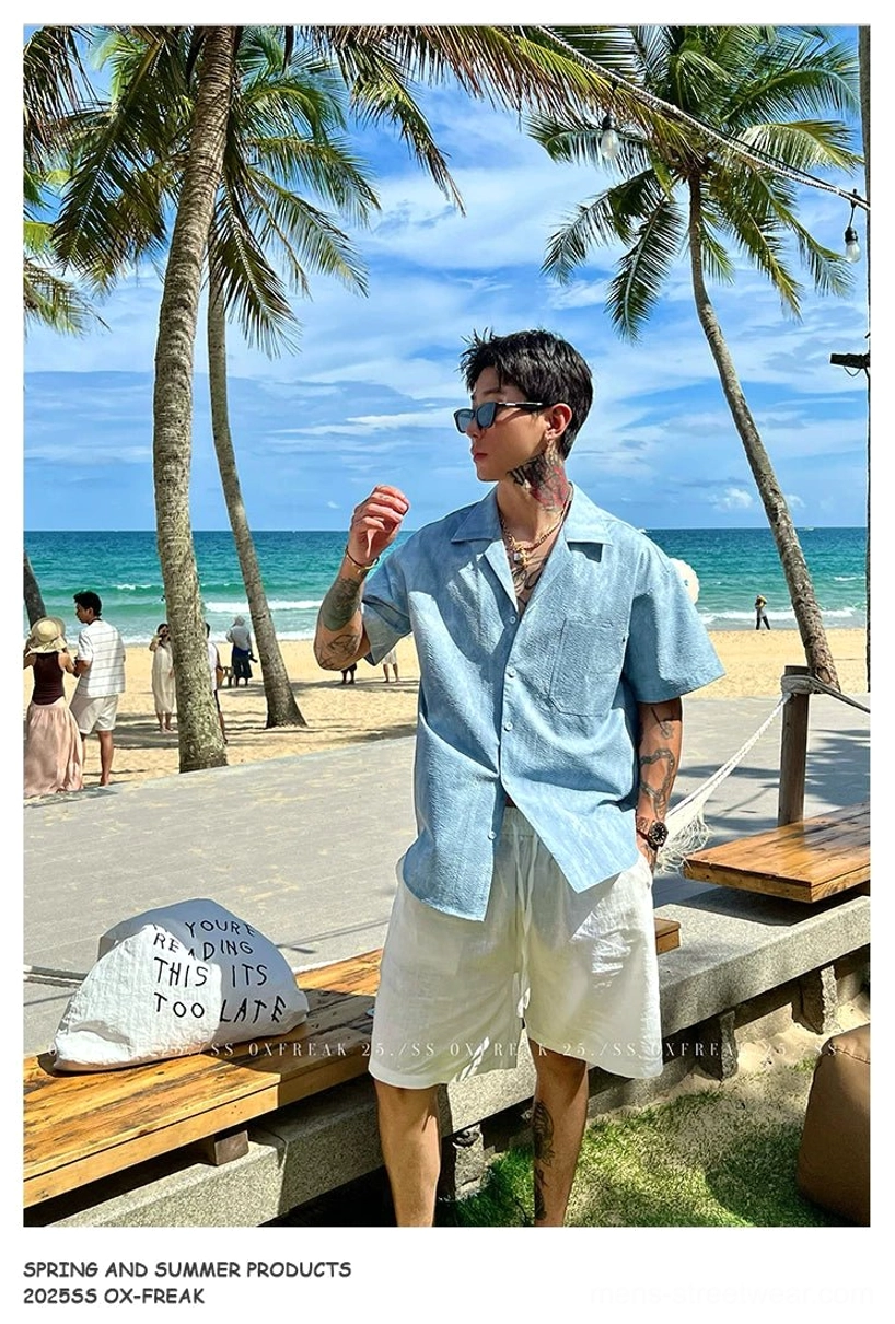 Blue washed Cuban collar short sleeve streetwear shirt styled with sunglasses beside a palm-lined seaside plaza