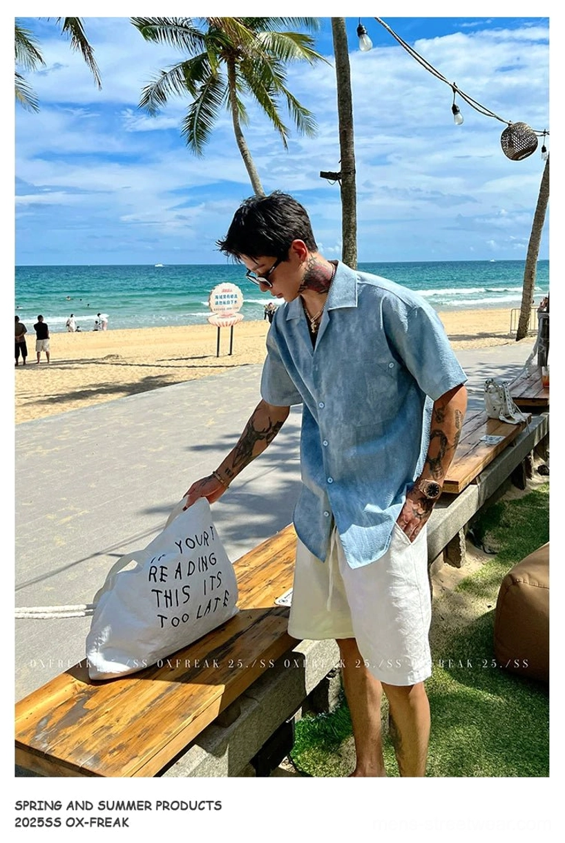 Blue wrinkled seersucker Cuban collar shirt styled with white shorts and a canvas tote near a seaside bench