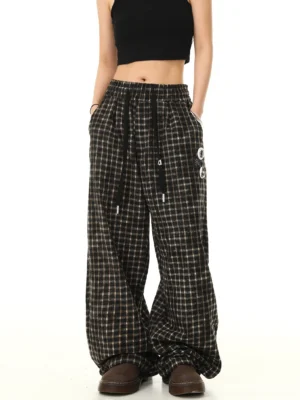 Front view of brown black check print wide leg track pants with graphic leg badges worn with a black crop top and platform shoes