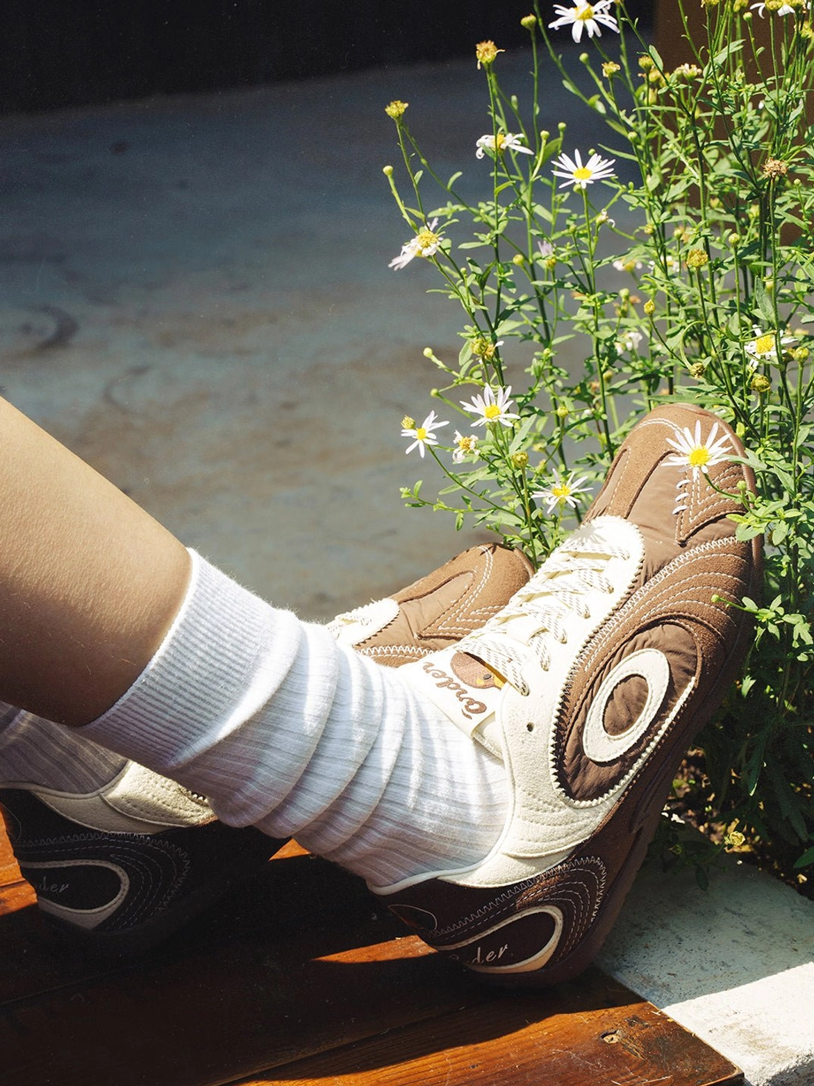 Brown retro streetwear trainers worn with white crew socks in a casual outdoor setting with daisy flowers