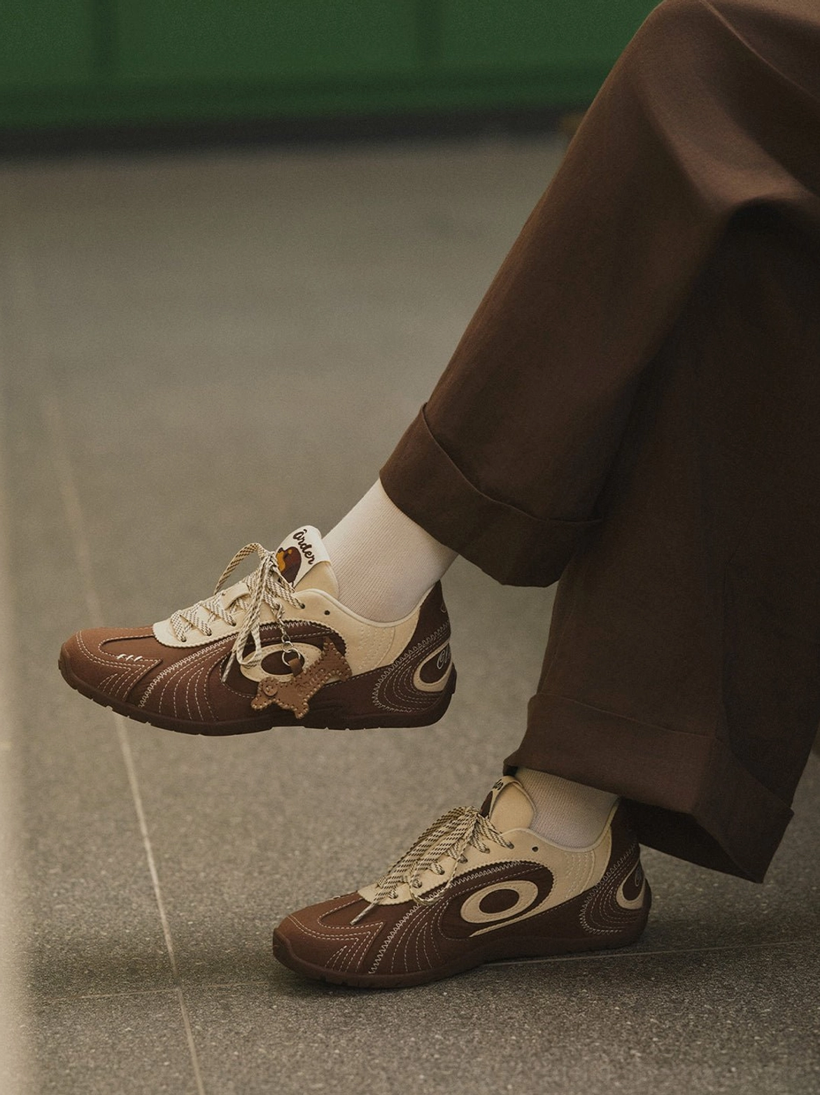 Brown retro low-top trainer showing the detachable dog pendant and tonal gum outsole in a close side view