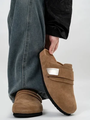 Brown soft suede strap mule clogs worn with loose blue jeans and a black quilted jacket, showing the rounded toe, adjustable fastening strap, metal accent plate, and thick rubber outsole