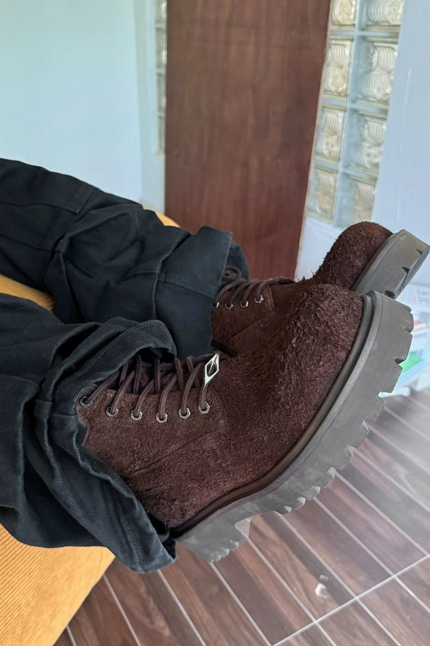 Side three-quarter view of brown suede combat boots with chunky gray lug soles and metal lace hardware, styled with black cargo pants