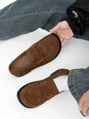 Soft suede penny loafer held above a casual outfit, showing the matte brown finish, decorative stitching, leather interior, and classic slip-on design