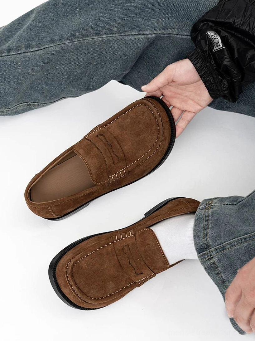 Soft suede penny loafer held above a casual outfit, showing the matte brown finish, decorative stitching, leather interior, and classic slip-on design
