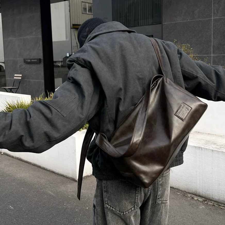 Brown waxed-look PU leather crossbody commuter bag worn over the shoulder with a dark gray utility jacket and jeans