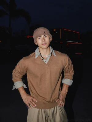 Camel brown V-neck knit sweater styled with a brown beanie, necklace, rolled checked shirt cuffs, and urban Seoul-inspired night streetwear