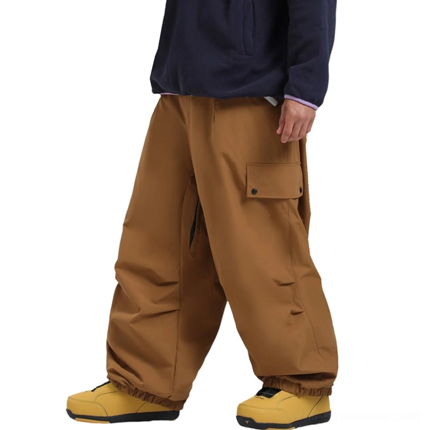Caramel brown loose-fit ski cargo pants shown from the side with elasticated cuffs, thigh flap pocket, wide-leg outdoor silhouette, and relaxed snowboarding style