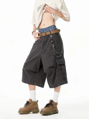 Charcoal gray oversized cargo Bermuda shorts shown from a low angle with wide baggy legs, large side pockets, cotton canvas texture, cropped streetwear tee, chain accessory, white socks, and work boots
