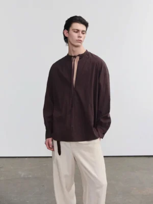 Chestnut brown men's oversized tie-neck long sleeve shirt with dropped shoulders styled with wide-leg trousers in a minimalist studio