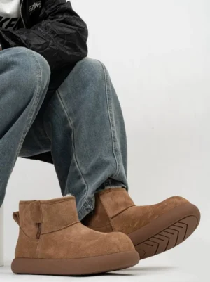 Chestnut brown men's shearling ankle boots worn with blue jeans and a black jacket, highlighting the rounded toe, suede upper, thick rubber sole, and side strap detail
