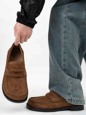 Chestnut brown suede penny loafer worn with baggy blue jeans and a black jacket, showing the square toe and low rubber sole in a modern streetwear outfit