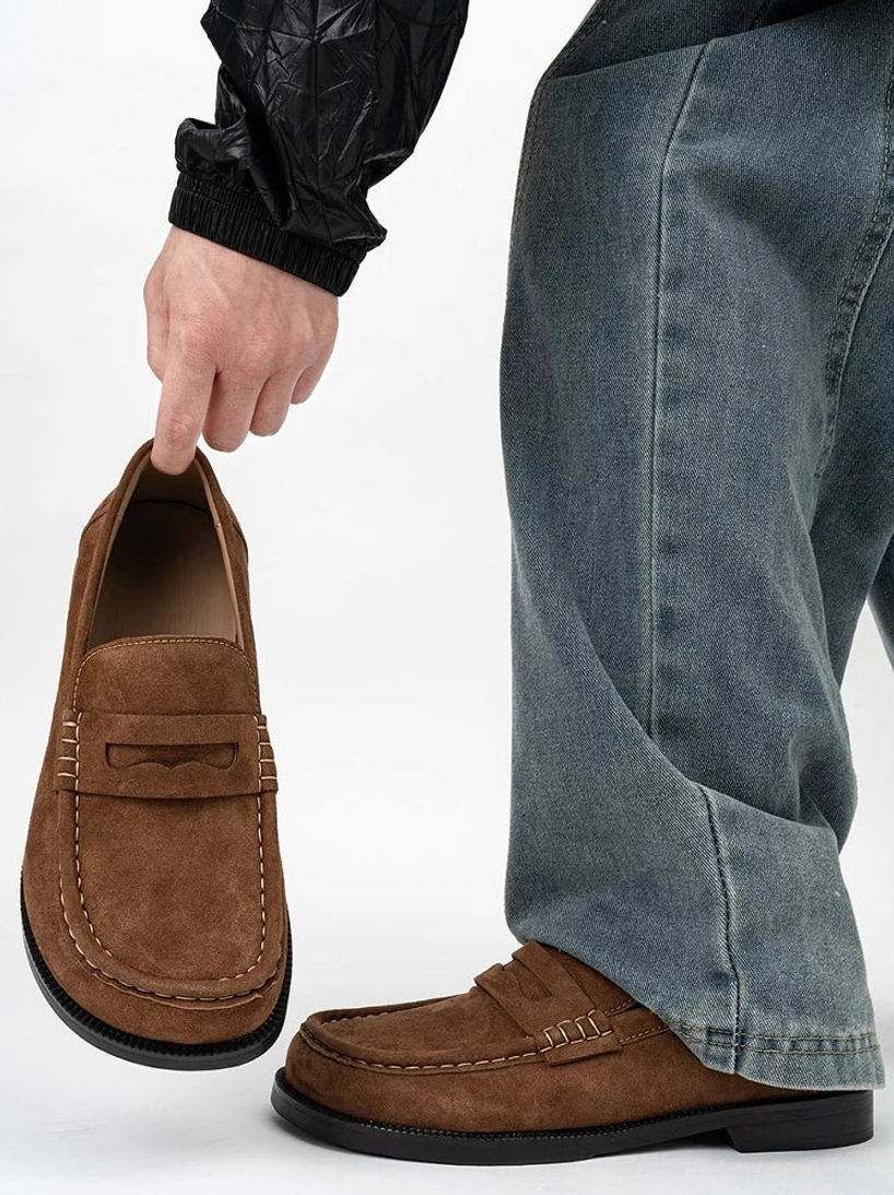 Chestnut brown suede penny loafer worn with baggy blue jeans and a black jacket, showing the square toe and low rubber sole in a modern streetwear outfit