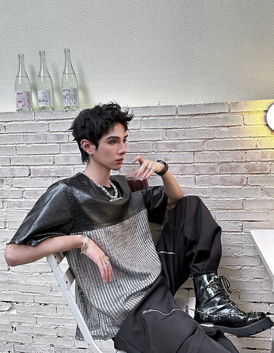Seated view of a black and silver chrome grid patchwork streetwear T-shirt with crocodile-effect upper panels, metallic texture, cargo pants, and boots