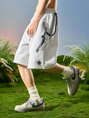 Side low angle view of light gray clover embroidered lounge sweat shorts with relaxed Korean streetwear fit, cotton blend fabric, rope drawstring waistband, and male model outdoors