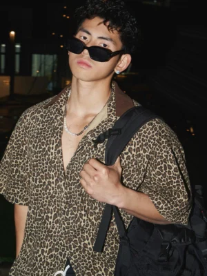 Close-up of a model wearing a coffee brown leopard print short-sleeve camp collar shirt with sunglasses, silver necklace, and black backpack in a Korean streetwear outfit