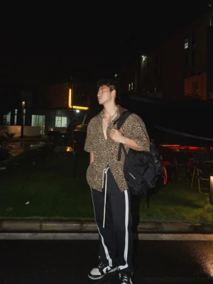 Side view of a relaxed leopard print short-sleeve button-up shirt styled with striped track pants, sneakers, and a black backpack