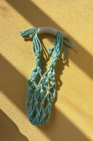 Light cyan colorful macrame water bottle carrier with white wrapped tassel handle shown empty on a yellow background