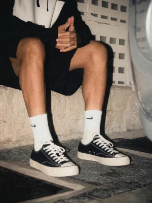 Male model wearing black cork canvas sneakers with white crew socks sitting on outdoor stone steps beside car streetwear styling